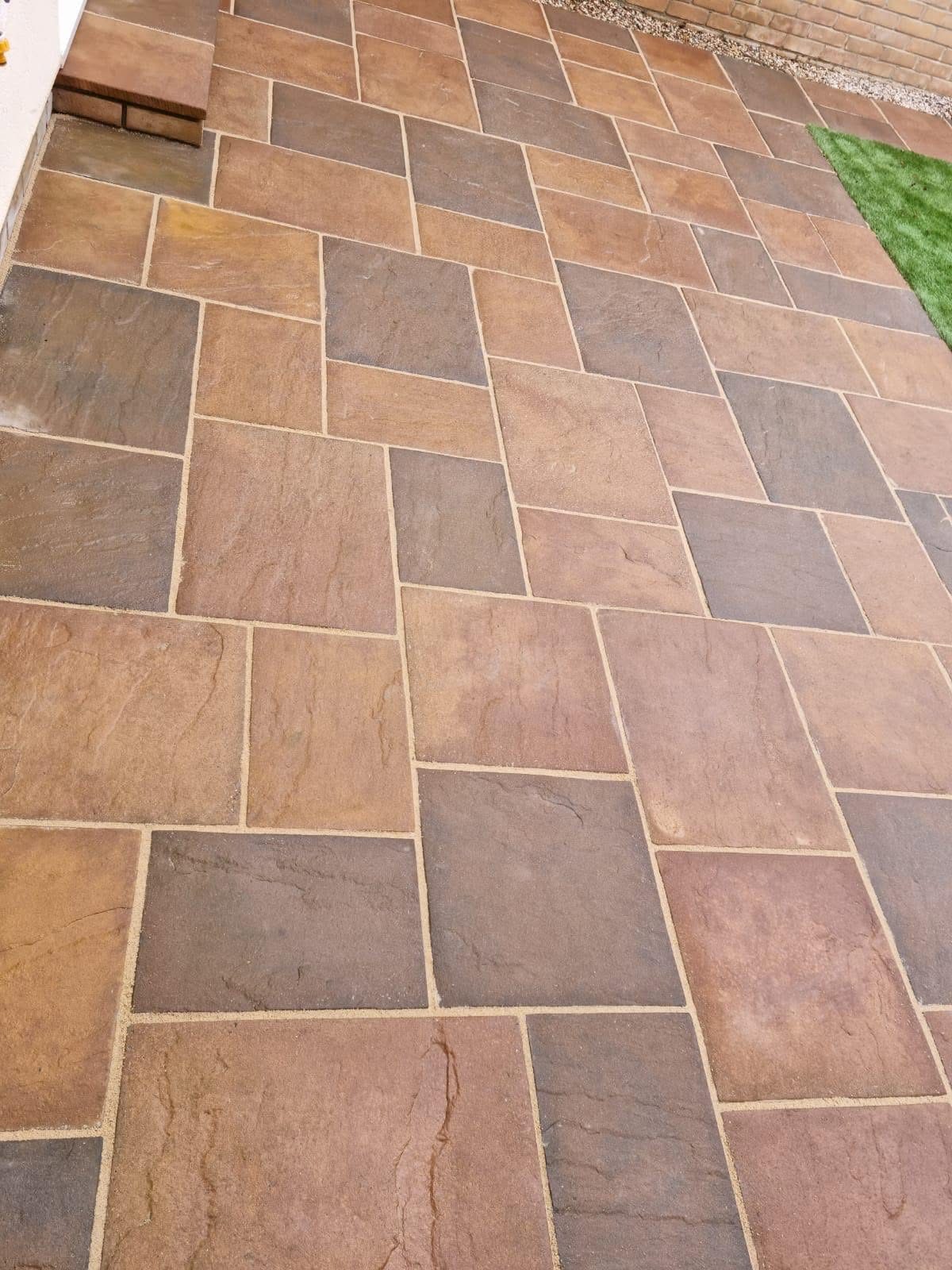 Close-up of a newly laid riven paving patio in warm autumn tones, tightly jointed and bordered by artificial grass